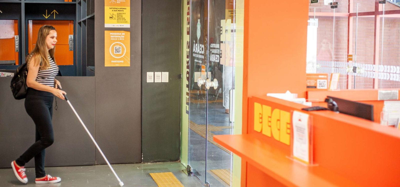A imagem mostra uma mulher com deficiência visual caminhando dentro da Biblioteca Pública Estadual do Ceará (Bece). Ela usa uma bengala branca para se orientar pelo piso tátil amarelo, que conduz à entrada de vidro do espaço. A mulher veste uma blusa listrada, calça preta, tênis vermelhos e carrega uma mochila preta. O ambiente apresenta tons de laranja e preto, com placas informativas sobre acessibilidade e sinalização tátil no chão. No balcão da recepção, destacam-se as letras amarelas formando "BECE". Ao fundo, a porta de vidro reflete parte do interior da biblioteca, e há sinais de saída visíveis.